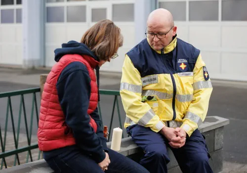 Ein Mitarbeiter der Notfallseelsorge sitzt auf einer Bank neben einer Frau. Zwischen Ihnen befindet sich eine angezündete Kerze